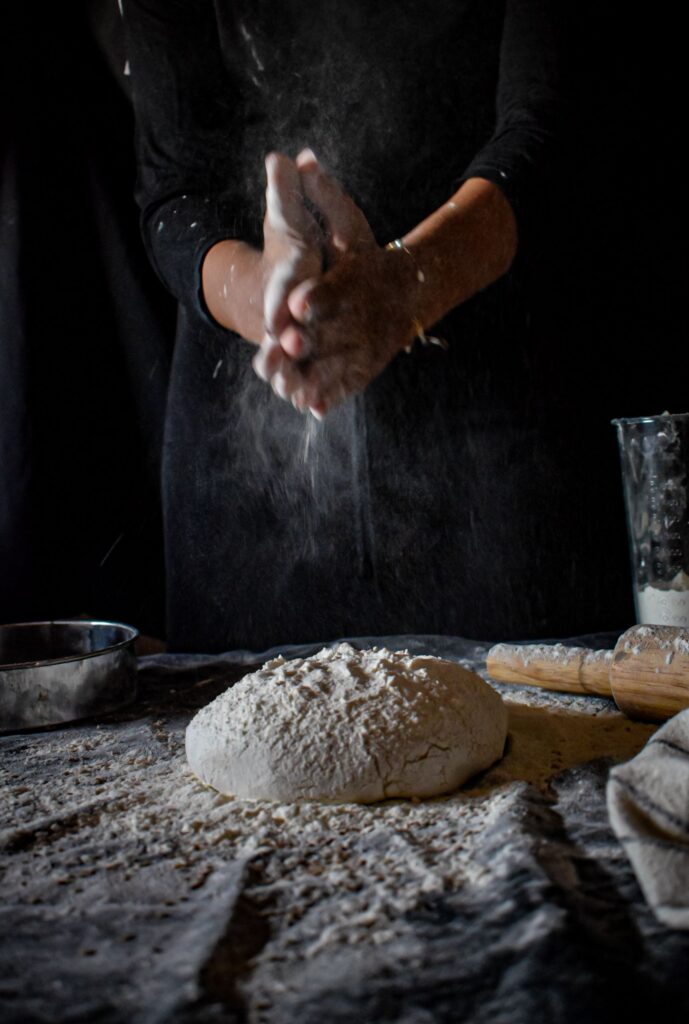 Close-up of hands dusting flour over dough on dark background, depicting baking process.